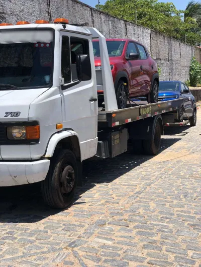Guincho com um carro vermelho na plataforma e um carro azul rebocado atrás, em rua de paralelepípedo